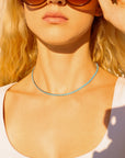 A woman in a white top and large brown sunglasses wears the Faire Turquoise Tennis Necklace, crafted from hypoallergenic 316L stainless steel, featuring sparkling rhinestones, with her wavy blonde hair glowing in sunlight.