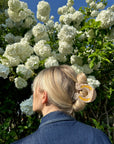 A person with blonde hair wears the Faire Fashion Hair Clip, crafted from eco-friendly cellulose acetate, while standing outdoors in a denim jacket near white flowering bushes under a clear blue sky.