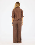 A person with long blonde hair stands facing away in the Kerri Rosenthal Tia Love Brigade Shirt—a relaxed button-down brown top with rolled sleeves—paired with matching wide-leg pants, set against a plain white background.