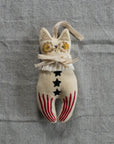 Introducing the "Rocket Cat Elton John" by Faire: a beautifully handmade fabric cat ornament, featuring gold-sequined eyes and a ribbon collar. This exquisite piece is filled with cotton and adorned with intricately embroidered stars and red stripes on its body, all set against a textured gray background.