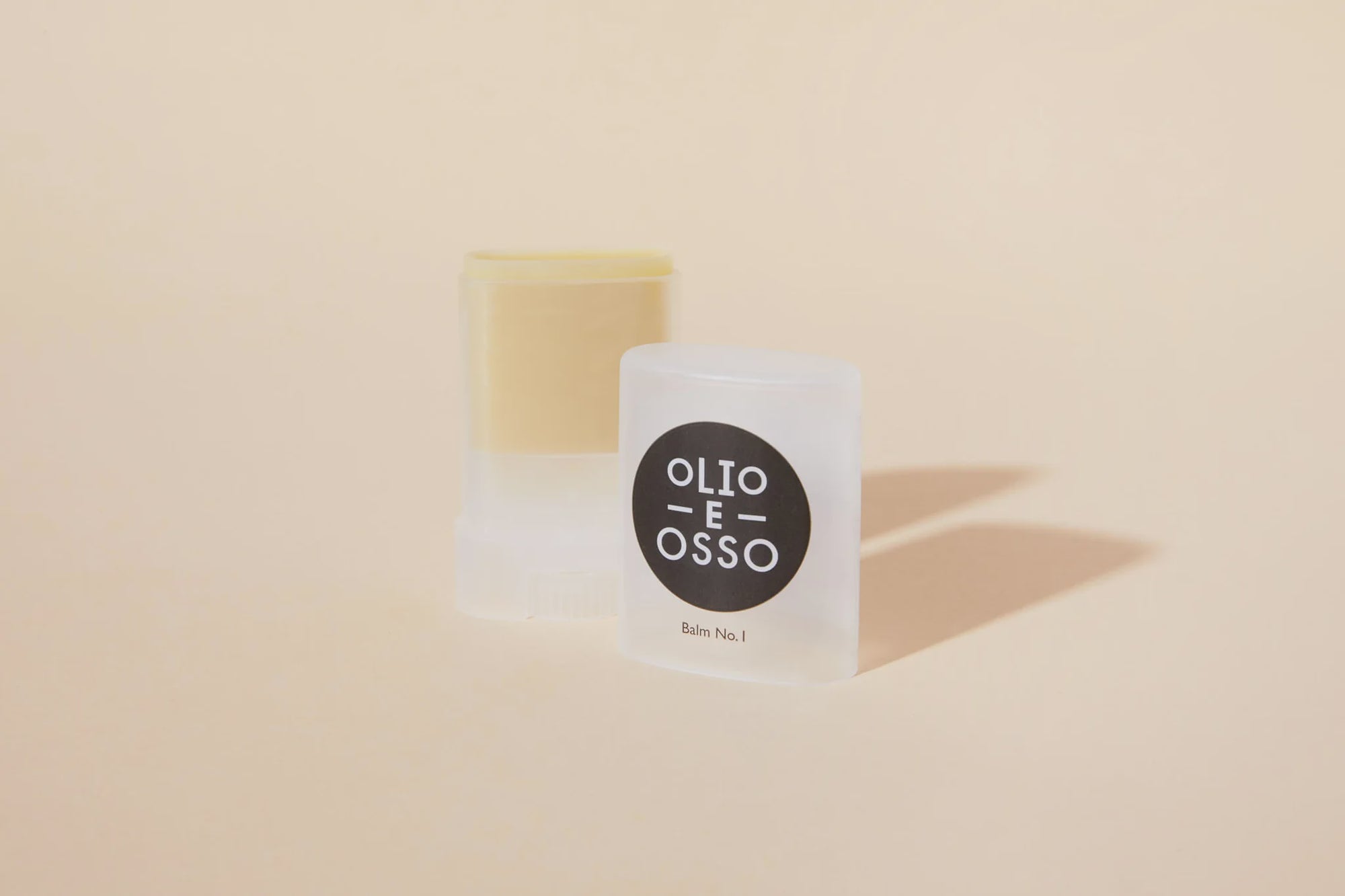 Two containers of Olio E Osso balm/stick, one opened with a yellow balm visible and one closed, displayed against a soft Arizona-style background.