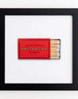 A framed piece featuring a red matchbox with the words "Restaurant Balthazar" printed on it. The matchbox contains several wooden matches with white tips. Encased in clear acrylic, the Match South Art Square Blk Frame contrasts beautifully with the white background, highlighting the matchbox.