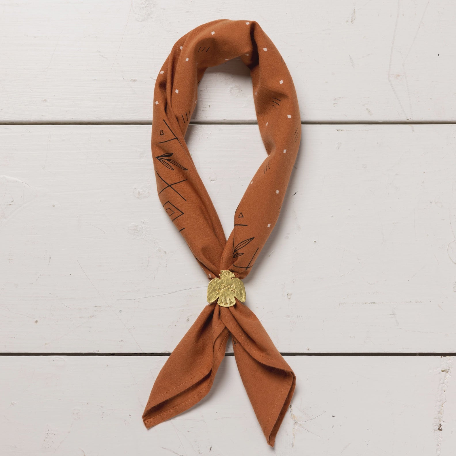 A brown silk scarf with black geometric patterns, elegantly tied and secured with a golden tassel, displayed on a white wooden background in Arizona style.