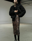 A person wearing a textured black faux fur jacket, the Le Superbe Liza Skirt, black lace-up boots, and a black beanie stands in a minimalist industrial-style space.