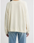 A person is shown from the back against a plain white background, wearing The Great Inc.'s The Rehearsal Sweatshirt in cream-colored cotton paired with light blue jeans.