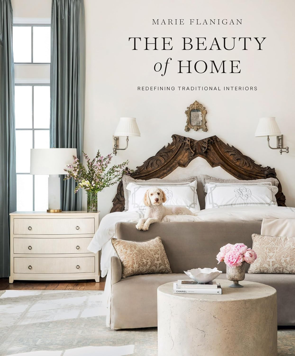 Elegant bedroom interior featuring a large bed with neutral bedding, an ornate wooden headboard, a side table with a lamp in Arizona style, and a dog sitting on the floor. Light decor with a title overlay "The Beauty of Home" by Gibbs Smith Publisher.
