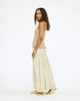 A woman with curly hair stands sideways against a plain white background, wearing a sleeveless beige top, gold flat shoes, and the cream-colored Kerri Rosenthal Vacance Pintuck Skirt.