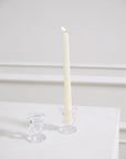 A tall, lit white candle in an urn-shaped clear Bellamy Candleholder by Accent Decor sits on a white surface beside an empty matching holder, all set against a white background with decorative wall molding.