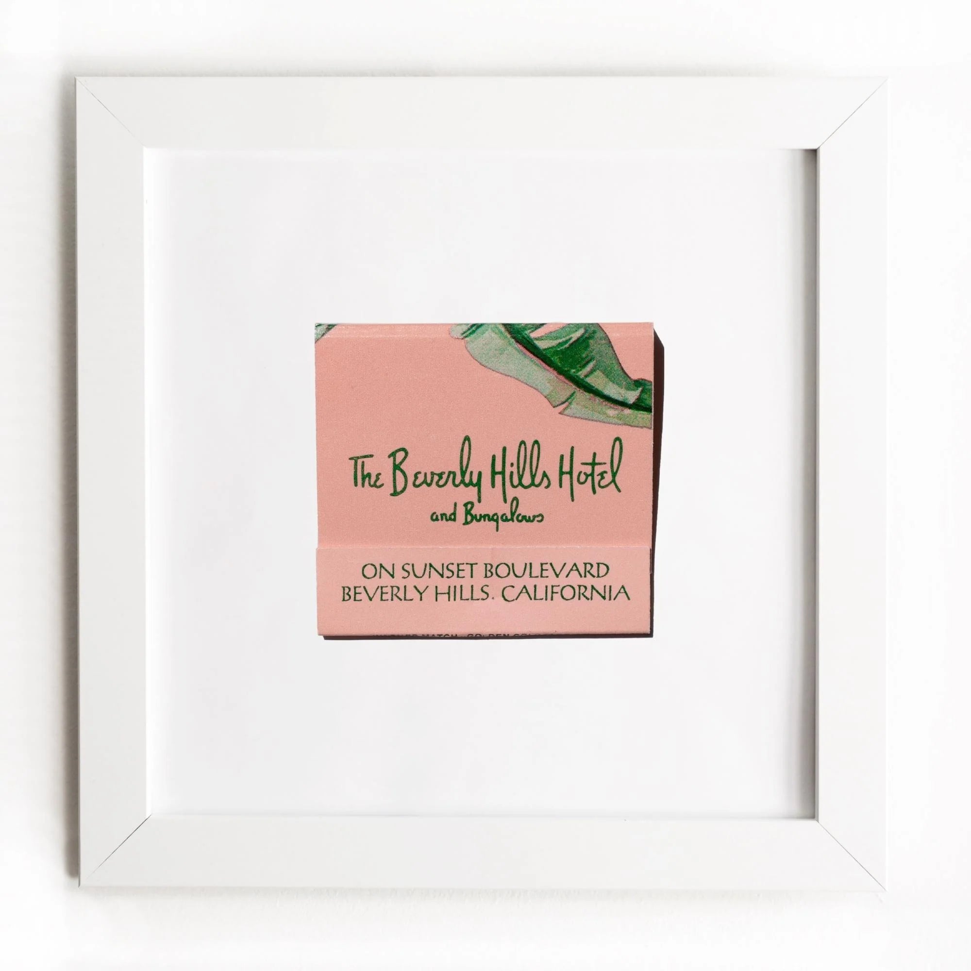 A framed artwork of a vintage hotel matchbook cover labeled "The Beverly Hills Hotel and Bungalows" in a Match South Art Square White Frame, featuring a pink background and green palm leaf design.