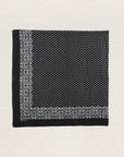 The Great Inc.'s The Big Bandana is a square black cotton scarf with a white floral border and dense white dots in the center, vintage-inspired and displayed flat on a neutral textured background.