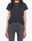 A woman wears Mother’s The Lil Goodie Goodie Tee in plain black, tucked into faded black jeans with layered gold necklaces and bracelets, standing against a white background.
