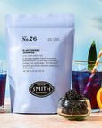 A bag of Faire Blackberry Jasmine Iced Tea stands next to a glass of iced tea with a striped straw, fresh blackberries, and an orange slice. A small bowl of blackberries is in the foreground.