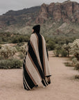 A person draped in the Faire TT Handwoven Blanket strolls along a desert path lined with cacti and shrubs, rocky mountains rising under a cloudy sky.