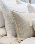 A neatly arranged bed adorned with a mix of textured pillows from POM POM's Lake Linen Collection, highlighted by the Blake Big Pillow 28x36 in beige with quilted tassels in the foreground. Smooth, white pillows featuring a sophisticated stripe pattern are positioned in the background, adding to the cozy and elegant atmosphere.