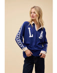 A blonde woman wears the DayDreamer Los Angeles Stars Rugby, an oversized blue raglan long sleeve with "LA" and white stars on the sleeves, paired with dark jeans against a beige background.
