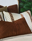 A bed with striped neutral pillows and a large Eastern Accents Rufus Nubuck Leather Pillow in front, set against a channeled headboard with a green leafy plant in the background.