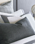 A neatly arranged bed features layered pillows in shades of gray and white, including the striking OCTAVIUS WOOL pillow by Eastern Accents, all set against a dark headboard with a blurred decorative sculpture in the background.