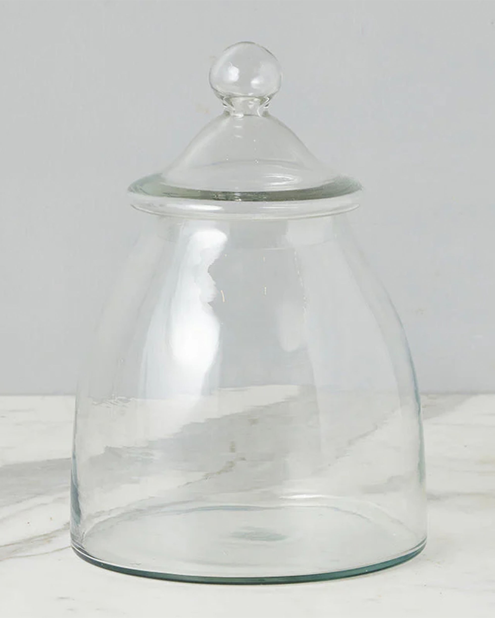 The Europe 2 You Glass Bon Bon Jar w/Lid features a clear, wide base with gently curved sides and a round-knob lid, displayed on white marble against a light gray background.