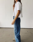 A person stands indoors against a white wall, wearing Hey Gang's The Boxy Polo in short sleeves and high-waisted blue jeans. Only part of their face is visible, with loose brown hair.