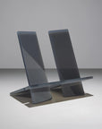 The Ingram Book Company’s Bookstand Md is a minimalist, modern chair crafted from translucent gray material with two angled backrests and a flat seat, set against a plain, light gray background.