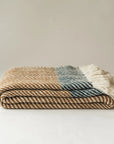 The Faire Chunky Twill Throw, featuring brown and cream patterns, dark blue checks, and white tassels on one end, is folded and displayed on a plain white surface.