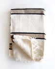 The TT Handwoven Blanket by Faire is a beige, textured, folded Mexican blanket with black and tan horizontal stripes and frayed edges, shown on a white surface.
