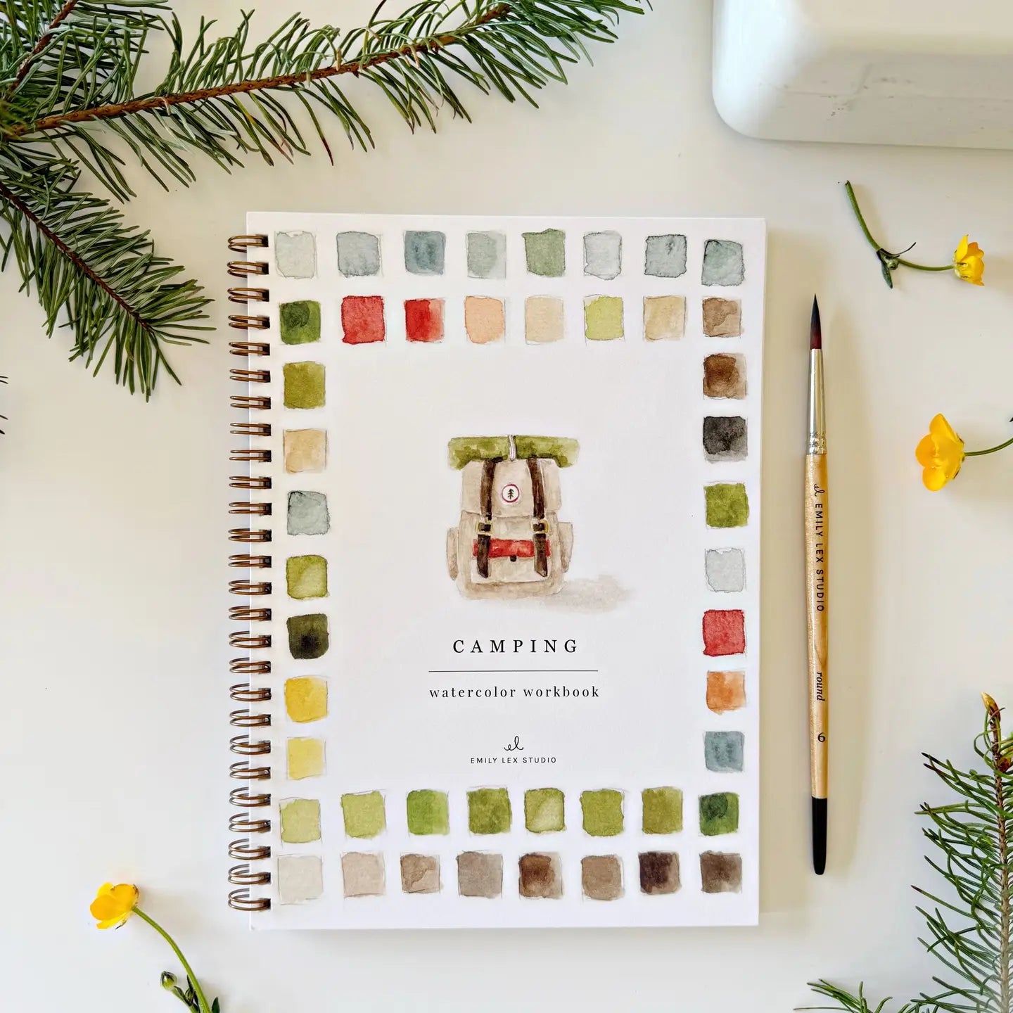 The Faire Watercolor Workbook is a spiral-bound notebook with watercolor squares and a painted camper on the cover. Beginner-friendly, it’s shown with fir branches, yellow flowers, and a paintbrush on a white surface.