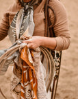 A person in a brown shirt with a patterned cowgirl accessory holds several ropes and colorful Wild Rag Silk Scarves by Faire, standing on sandy ground. Their face is not visible.
