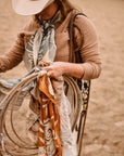 Wearing a wide-brimmed hat and the Faire Rustic Wild Rose Western Silk Scarf, a person stands on sandy ground holding several ropes and colorful bandanas, their face partially hidden by the hat.