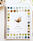 The Faire Watercolor Workbook for beginners, titled "Birds," is spiral-bound and features a painted bird cover with colorful paint swatches. It’s shown on a white surface beside a paintbrush and leafy plant.