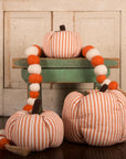 Three Faire Pumpkin decor pieces with orange and white stripes are paired with a felt ball garland on a small green bench, creating charming farmhouse style against a rustic wood background.
