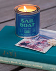 A lit G&W Soy Candle by Faire from the Happy Place Collection sits on two stacked books, including "Expert Sailing" by Bill Robinson, beside an old seaside photo and a seashell, evoking nostalgic joy.