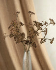 The Indaba Meadow Grass Bouquet, with dried brown stems and clustered flowers, sits in a clear glass vase against a beige fabric and off-white wall—offering natural faux botanical decor.