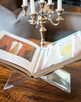 An open coffee table book is showcased on the Faire Acrylic Book Stand Display atop a wooden table next to a silver candelabra, with a blurred pillow and plaid furniture in the background.