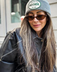 A woman with long, light brown hair smiles softly while wearing the Faire Patch Beanie Strong as a Mother in gray, paired with brown sunglasses and a black leather jacket outdoors in front of a building.