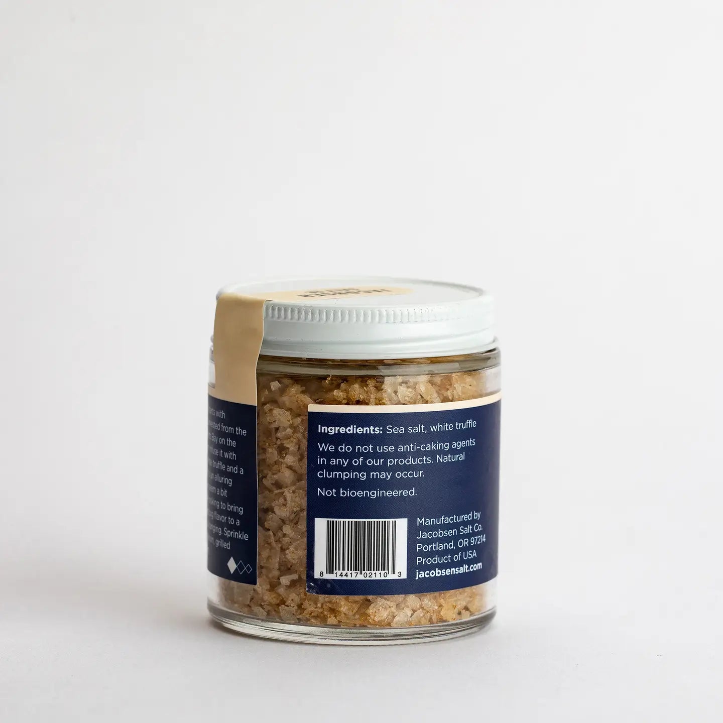 A small glass jar with a white lid holds Faire's White Truffle Salt, a coarse, light brown blend. The label lists "Ingredients: Sea salt, white truffle" and provides product details plus the manufacturer's address.