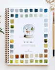 The spiral-bound Faire Watercolor Workbook, titled "Winter," showcases a snow globe with a deer, trees, and color swatches on the cover. Beside it are a pine sprig, paintbrush, and snowflake ornament set against a white background.