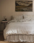 A neatly made bed features the Faire Kemptown Stripe Linen Duvet against a beige paneled wall. A landscape painting hangs above, while a bedside table holds books, a lamp, and decorative decor.