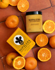 An overhead view of Faire Tangerine Cactus Body Polish in a jar with its matching yellow box, surrounded by whole and halved oranges on a terracotta surface.