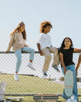 Five kids hang out by a chain-link fence on a sunny day. Two sit atop the fence while three stand or lean nearby, showing off their Faire Vintage Skateboards. All are dressed casually and enjoying time together.