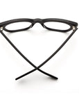 A pair of Caddis Miklos Matte Black eyeglasses is placed upside down on a white surface, with the arms open and crossed in a V shape.