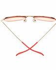 Caddis Mabuhay Polished Gold aviator sunglasses with gold wire frames, pink-tinted lenses, and red rubber temple tips are displayed upright with crossed arms on a white background.