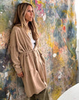 A woman with long hair wears the Kerri Rosenthal Cali Fringe Cardigan Coat, paired with green pants, standing against a colorful, abstract painted backdrop and gazing thoughtfully to the side.