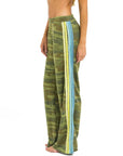 A person wears Aviator Nation's 5 stripe women's wide leg pocket sweatpants—vintage, loose-fitting green camo with blue and yellow vertical side stripes—standing barefoot against a plain white background.