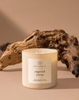 A white Hico Candle from Faire sits before textured driftwood, its Cattleman fragrance adding freshness to the beige background and soft shadows.