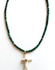 The Carmelia Necklace by Faire features alternating dark green malachite cubes and gold beads, accented with a white cross pendant adorned with a small gold heart at the center, displayed on a plain white background.