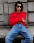 A person with short dark hair, black sunglasses, and blue jeans sits on a stone bench against a gray block wall, arms folded and legs crossed, wearing a bright red Sold Out NYC Cashmere Polo.