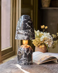 The Caswell Table Lamp by Abigail Ahern, featuring a black and white marble finish, sits on a wooden desk near an open book and dried flowers, providing stylish accent lighting beside a sunlit window.