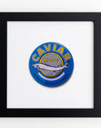 Framed artwork featuring a circular design with a sturgeon fish illustration in the center and the word "CAVIAR" in bold yellow letters. The background is blue with additional text and details in smaller fonts below the fish. This exquisite piece, Match South's Art Square Blk Frame, is protected by clear acrylic, enhancing its modern appeal.
