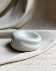 The Sumo Pocket Concrete Bowl by Faire, featuring a small, smooth, rounded and irregular design, rests on a textured stone surface in wabi sabi style with draped beige fabric in the background.
